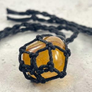 Pode incluir: Uma gema amarela envolta em uma rede de macramê preta. A gema é um cristal natural com textura áspera. A rede de macramê é feita à mão e tem um design delicado.