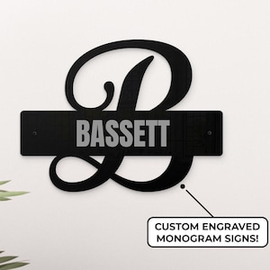 May include: Black metal monogram sign featuring the letter B and the name "BASSETT" in a silver font. The sign is mounted on a white wall. "CUSTOM ENGRAVED MONOGRAM SIGNS!" is written in a white box below the sign.