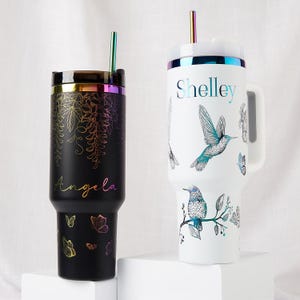 Könnte beinhalten: Zwei isolierte Becher mit schillernden Akzenten und Strohhalmen. Einer ist schwarz mit einem Blumendesign und dem Namen "Angela". Der andere ist weiß, mit Kolibris und Schmetterlingen, mit dem Namen "Shelley". Beide haben Deckel.