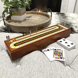 May include: A wooden cribbage board with yellow, black, and white markings, pegs, and a deck of playing cards. The board is brown and rectangular, with a curved track. The cards show various suits and face cards.