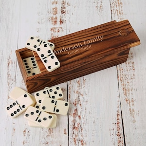 May include: A wooden domino set with a dark stain, featuring the engraved text "Anderson Family Game Night." The set includes ivory-coloured dominoes with black dots, some inside the wooden box and others scattered around.