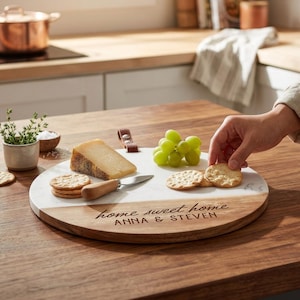 May include: A round serving board made of white marble and wood, featuring the engraved text "home sweet home" and "ANNA & STEVEN". The board includes a leather handle loop and is arranged with a wedge of cheese, green grapes, and crackers.
