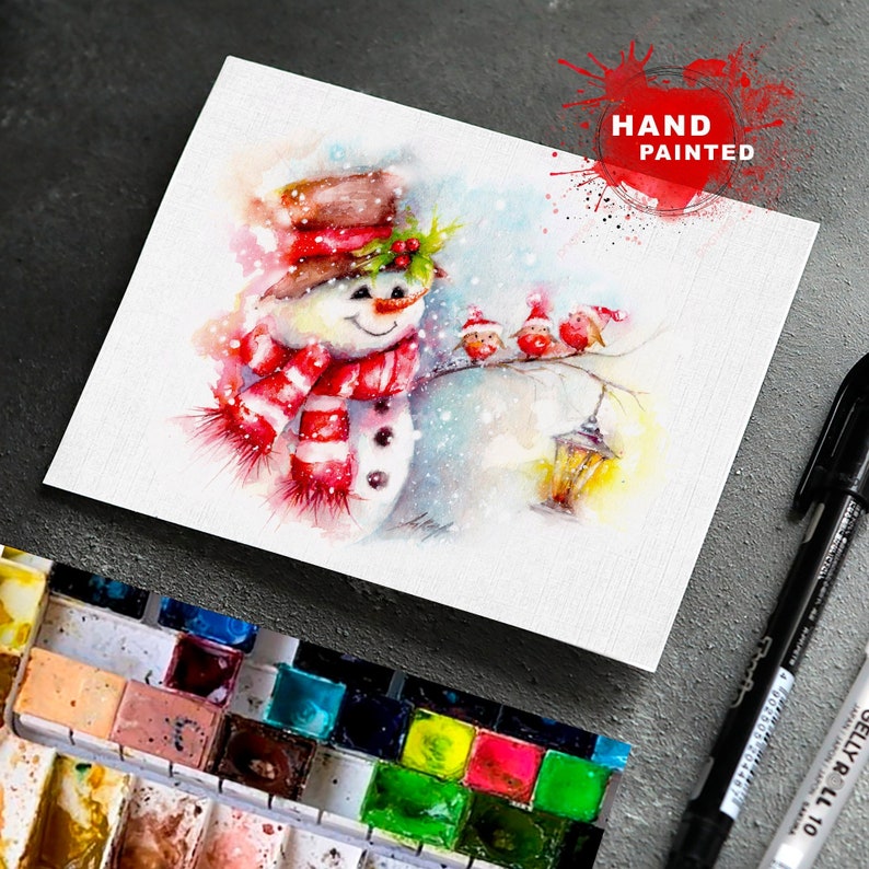 May include: Watercolour painting of a snowman wearing a red and white striped scarf and a brown hat with holly berries. Three red birds are perched on a branch with a lantern hanging nearby.