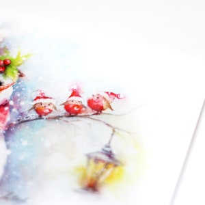 May include: Watercolour painting of a snowman wearing a red scarf and hat, standing in the snow with three red robins wearing Santa hats perched on a branch.