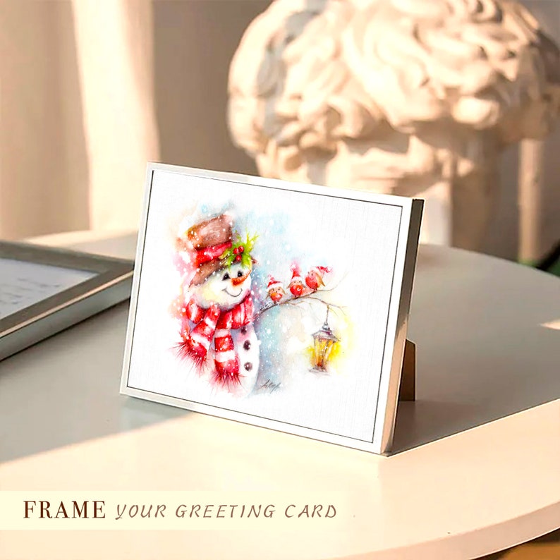 May include: A white framed greeting card featuring a watercolour illustration of a snowman wearing a red and white scarf and hat. Three red birds are perched on a branch above the snowman, and a lantern hangs from the branch. The card is on a white surface.