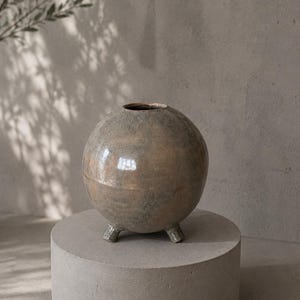 May include: A spherical ceramic vase with a mottled, earthy brown and grey glaze finish. The vessel features a small circular opening at the top and is supported by three short, sturdy feet at the base, creating a balanced and rounded silhouette.