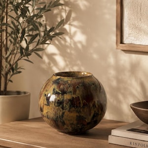 Könnte beinhalten: Eine kugelförmige Keramikvase mit einer glänzenden, mehrfarbigen Glasur in gemischten Tönen von Dunkelbraun, Ocker und Dunkelgrün. Die Oberfläche weist ein gesprenkeltes, tropfenartiges Muster mit einem glatten, reflektierenden Finish auf.