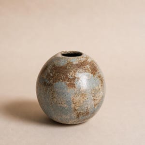 Könnte beinhalten: Eine kugelförmige Keramikvase mit einer kleinen kreisförmigen Öffnung an der Oberseite. Die Oberfläche weist ein gesprenkeltes, strukturiertes Finish mit einer Mischung aus gedeckten blaugrauen und erdigen Brauntönen auf.