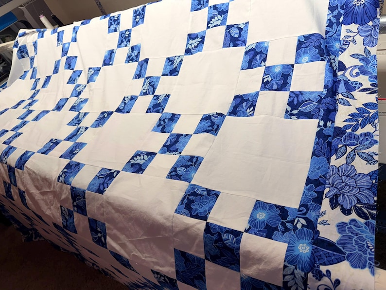 K&ouml;nnte beinhalten: Eine handgefertigte Steppdecke mit einem wei&szlig;en und blauen Blumenmuster. Die Steppdecke hat ein Schachbrettmuster mit wei&szlig;en Quadraten und blauen Blumenquadraten. Der Rand ist ein blaues Blumenmuster. Die Steppdecke ist aus weichem Stoff.