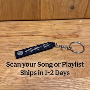 Könnte beinhalten: Schwarzer rechteckiger Schlüsselanhänger mit einem Spotify-Code und einem silbernen Schlüsselring. Der Text "Scan your Song or Playlist" ist unterhalb des Schlüsselanhängers aufgedruckt. Der Schlüsselanhänger liegt auf einer Holzoberfläche.