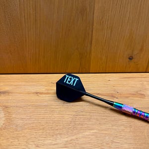 May include: A black dart with the word "TEXT" in white on the flight. The dart has a black shaft and a colorful, iridescent grip. The dart is resting on a wooden surface.