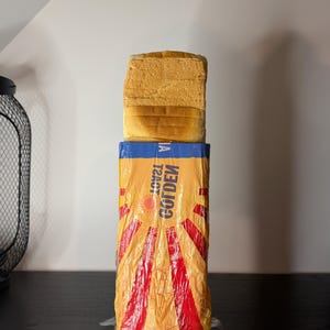 Pode incluir: Um pão dourado fatiado está em cima de um saco plástico amarelo e vermelho. O saco tem a palavra "GOLDEN" e um desenho de raios de sol. O pão é castanho claro e parece fresco.