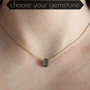 Op de afbeelding: Een fijne gouden ketting met een gefacetteerde, iriserende edelsteen hanger. De ketting wordt gedragen, met de ketting zichtbaar op de huid. Tekst bovenaan luidt "choose your gemstone".