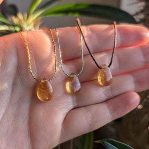 May include: Three necklaces featuring teardrop-shaped yellow citrine pendants. The first pendant hangs on a gold-toned chain, the second on a silver-toned chain, and the third on a dark brown cord. Each gemstone is translucent and faceted.