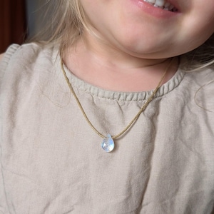 Op de afbeelding: Een kind draagt een lichtbruin shirt met een eenvoudige, traanvormige ketting. De hanger is gemaakt van iriserend glas en hangt aan een bruin koord.