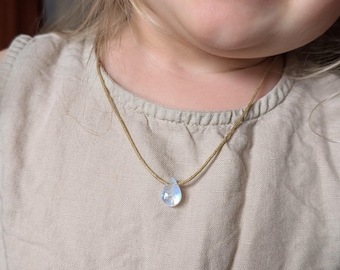 Peut inclure: Un pendentif en forme de goutte avec une finition translucide et irisée, suspendu à un cordon fin de couleur beige. Le pendentif reflète la lumière avec des tons bleus et blancs subtils sur un vêtement en tissu beige texturé.