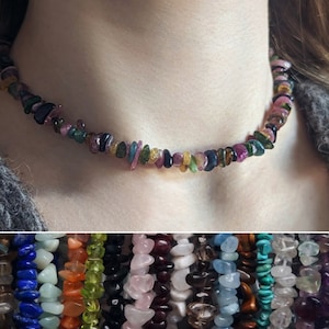 May include: A multi-coloured gemstone necklace made of small, irregular shaped chips. The necklace is made of various colours including pink, green, blue, black, and yellow. The necklace is worn by a person with long brown hair.
