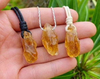 Puede incluir: Tres colgantes de cristal de citrino en bruto, cada uno con una piedra de color amarillo dorado translúcido. El colgante izquierdo cuelga de un cordón negro, el central de una cadena de metal plateado y el derecho de un cordón crema anudado.