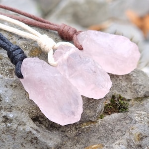 Può includere: Tre ciondoli in cristallo di quarzo rosa di colore rosa su cordini di diversi colori. I cordini sono marrone, bianco e nero.