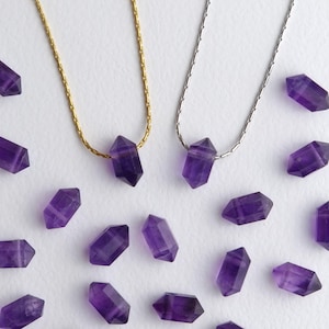 May include: Two necklaces with amethyst crystal pendants, one on a gold chain and the other on a silver chain. Several loose amethyst crystals are scattered around the necklaces on a white surface. The crystals are a deep purple color.