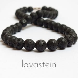 Puede incluir: Un collar compuesto por cuentas redondas y porosas de piedra de lava negra dispuestas en un bucle continuo. Las cuentas presentan una superficie rugosa y texturizada característica de la roca volcánica. La palabra lavastein aparece debajo.