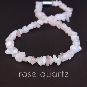 May include: A rose quartz necklace made of small, irregular, pale pink and white stones. The necklace is strung together with a silver-coloured clasp. The words "rose quartz" are written in white script below the necklace against a dark background.