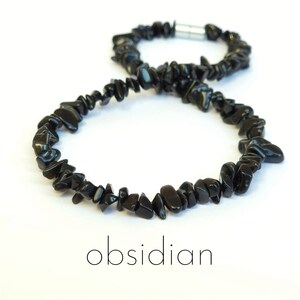 Peut inclure: Un collier composé de nombreuses petites pépites de pierre d'obsidienne noire polie disposées en boucle continue. Le collier présente un fermoir cylindrique en métal argenté et le mot obsidian est imprimé en noir sous l'objet.