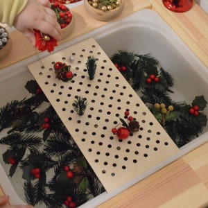 May include: A white plastic tub filled with water and green pine branches, red berries, and a wooden grid with holes. A hand is reaching into the tub to add more decorations.