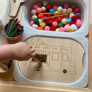 May include: A wooden ice cream van toy with a cutout window and a sign that reads "glace - gaufre - crepe - bonbon". The van is on a white surface with a bowl of colourful pom-poms in the background.