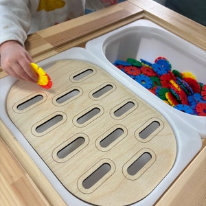 Può includere: Un tavolo da gioco in legno con un bacino di plastica bianco pieno di pezzi di plastica a forma di fiore colorati. Il tavolo ha una forma ovale ritagliata con più piccoli ritagli ovali. Una mano di bambino sta posizionando un pezzo a forma di fiore giallo e rosso in uno dei ritagli.