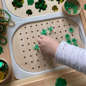 May include: A wooden board with a pegboard section and a cutout section. The pegboard is filled with green shamrock-shaped pegs. A child's hand is placing a peg into the board.
