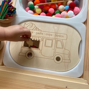 May include: A wooden toy food truck with cut-out windows and a sign that reads "gaufre - crepe - bonbon". A child's hand is placing a wooden food item into the truck.