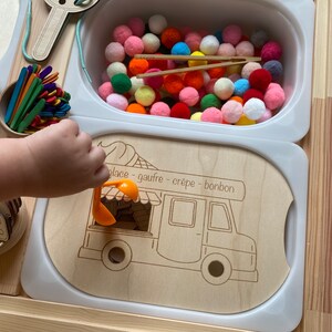 May include: A wooden toy food truck with a white background and a scoop in the window. The truck has the words "place - gaufre - crepe - bonbon" written on the side. There is a bowl of colourful pom-poms in the background.