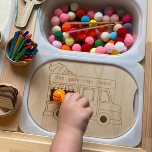 May include: A wooden toy ice cream van with a white background and the words "glace - gaufre - crepe - bonbon" written on it. A child's hand is reaching into the van to grab a small orange toy.