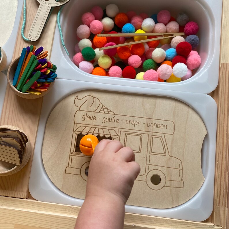 May include: A wooden toy ice cream van with a white background and the words "glace - gaufre - crepe - bonbon" written on it. A child's hand is reaching into the van to grab a small orange toy.
