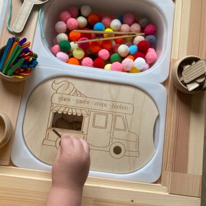 May include: A wooden toy ice cream van with a white background and the words "glace - gaufre - crepe - bonbon" written on the side. The van has a window with a small wooden ice cream cone inside. There are also colourful pom-poms in a white container.