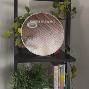May include: A round mirror with the text "SHORT N' SWEET" and "PARENTAL ADVISORY EXPLICIT CONTENT" is displayed on a black shelf. The shelf also holds a small potted plant, books, and a larger green plant. The mirror has a light brown wooden design.