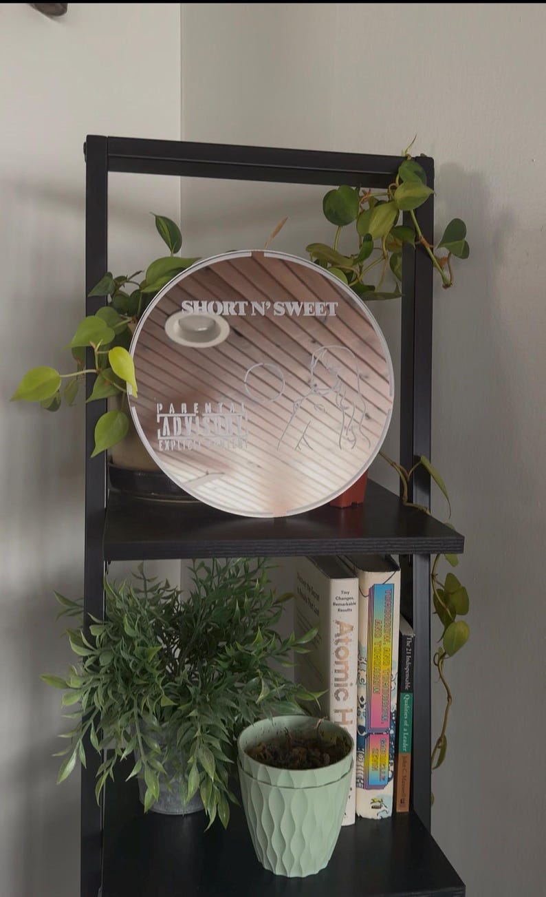 May include: A round mirror with the text "SHORT N' SWEET" and "PARENTAL ADVISORY EXPLICIT CONTENT" is displayed on a black shelf. The shelf also holds a small potted plant, books, and a larger green plant. The mirror has a light brown wooden design.