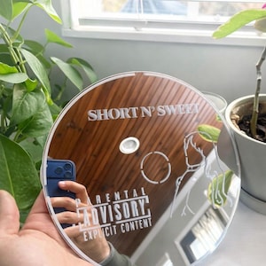 May include: A clear, circular mirror with etched designs and text. The text reads "SHORT N' SWEET" and "PARENTAL ADVISORY EXPLICIT CONTENT". The mirror reflects a hand holding a phone. The design includes a hand and a circle.