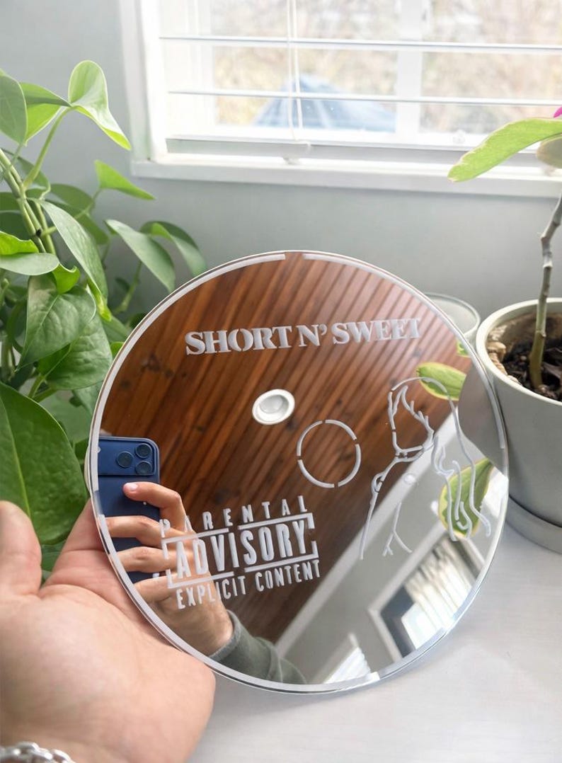 May include: A clear, circular mirror with etched designs and text. The text reads "SHORT N' SWEET" and "PARENTAL ADVISORY EXPLICIT CONTENT". The mirror reflects a hand holding a phone. The design includes a hand and a circle.