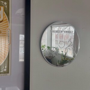 May include: A round, reflective mirror with the text "SHORT N' SWEET" and a female figure etched on its surface. The mirror is mounted on a light gray wall, with a framed print visible to the left.