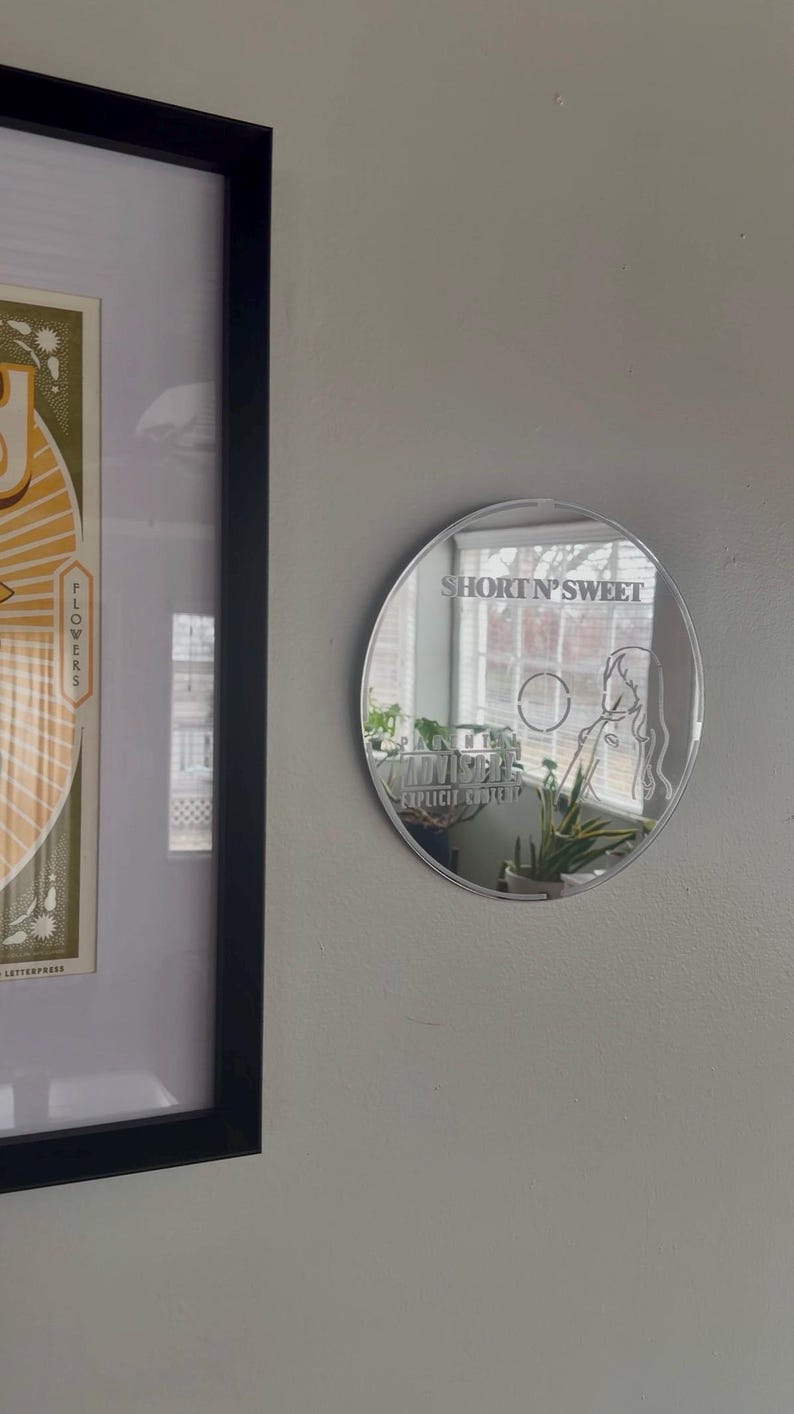 May include: A round, reflective mirror with the text "SHORT N' SWEET" and a female figure etched on its surface. The mirror is mounted on a light gray wall, with a framed print visible to the left.