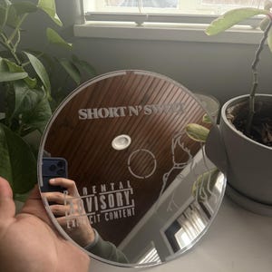 May include: A round, mirrored surface with the text "SHORT N' SWEET" and "RENTAL ADVISORY EXPLICIT CONTENT" etched into the surface. The mirror reflects a hand holding a phone and a room with plants.