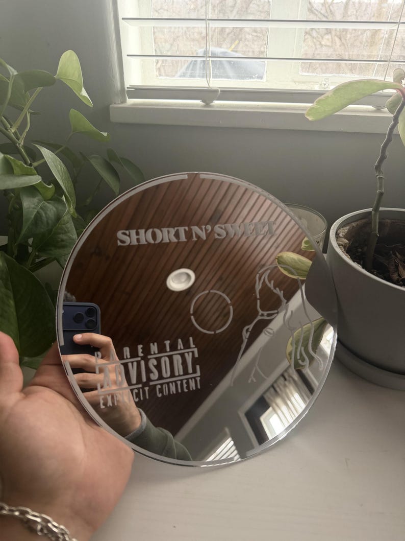 May include: A round, mirrored surface with the text "SHORT N' SWEET" and "RENTAL ADVISORY EXPLICIT CONTENT" etched into the surface. The mirror reflects a hand holding a phone and a room with plants.