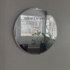 May include: A round mirror with the text "SHORT N' SWEET" at the top and "PARENTAL ADVISORY EXPLICIT CONTENT" at the bottom. The mirror reflects a room with a window and plants.