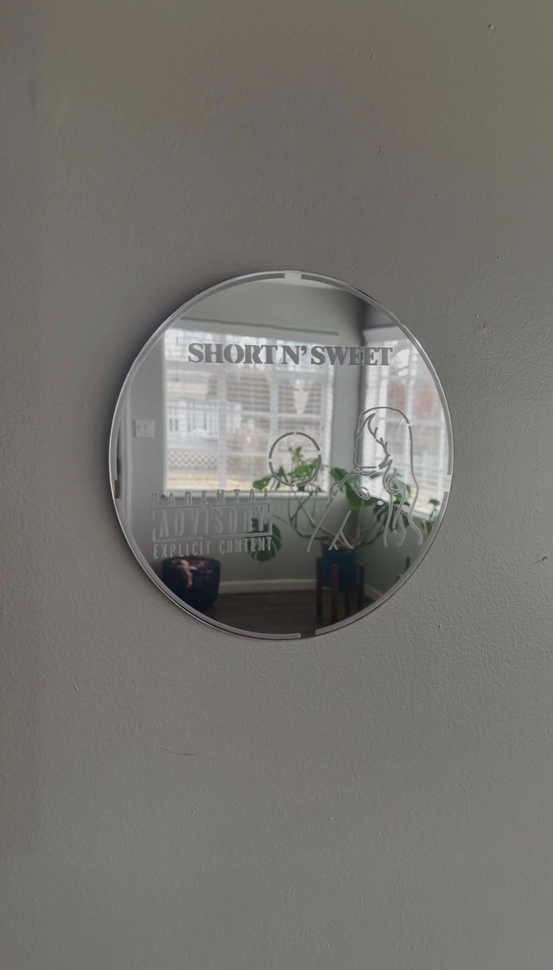 May include: A round mirror with the text "SHORT N' SWEET" at the top and "PARENTAL ADVISORY EXPLICIT CONTENT" at the bottom. The mirror reflects a room with a window and plants.