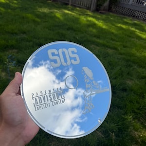 May include: A reflective CD with the text "SOS" and "Parental Advisory Explicit Content." The CD reflects a blue sky with clouds and a silhouetted figure. The disc is held in a hand.