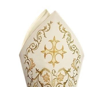 Może przedstawiać: Biała mitra biskupia z haftem złotym w kształcie krzyża i wzorów kwiatowych. Mitra ma białą wstążkę satynową z ozdobnymi złotymi paskami.