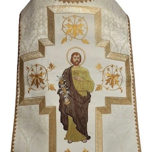 May include: White liturgical vestment with gold embroidery depicting a saint holding a flower. The embroidery features a cross with floral designs and a Greek key border.