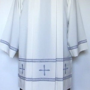 May include: White liturgical vestment with a black collar and blue embroidered crosses. The vestment has a pleated front and a gathered skirt.